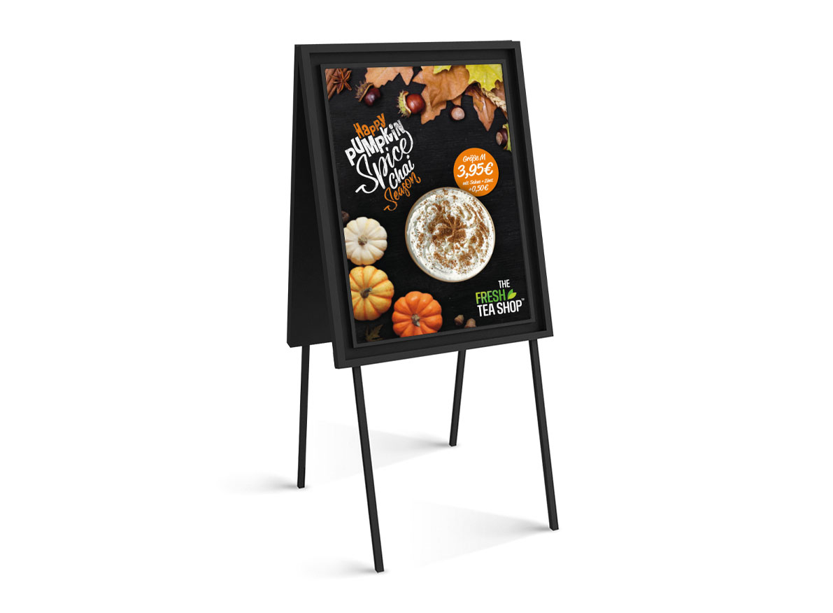 Auf einem schwarzen A-förmigen Schild ist eine herbstliche Werbung für „Vollblut The Fresh Tea Shop“ zu sehen. Darin sind ein Kürbisgewürzgetränk, Minikürbisse und Herbstblätter zu sehen. Der Preis beträgt 3,95 €, darunter ist das Logo des Ladens zu sehen.