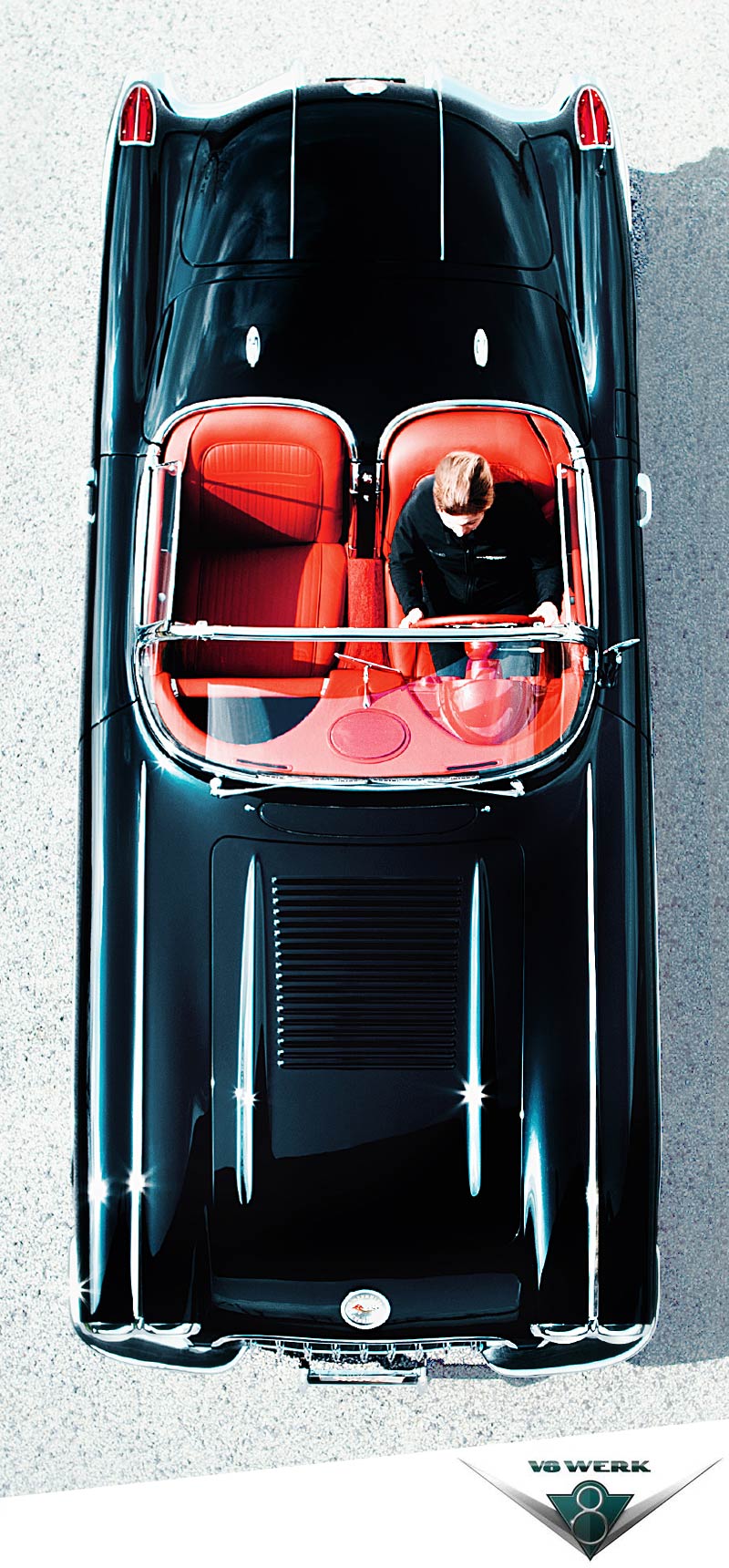 Draufsicht auf ein klassisches schwarzes Cabrio mit roten Sitzen, geparkt auf einem hellen Untergrund. Auf dem Fahrersitz sitzt eine Person mit hellem Haar. Am unteren Bildrand ist das Logo von Vollblut V8 Werk zu sehen.