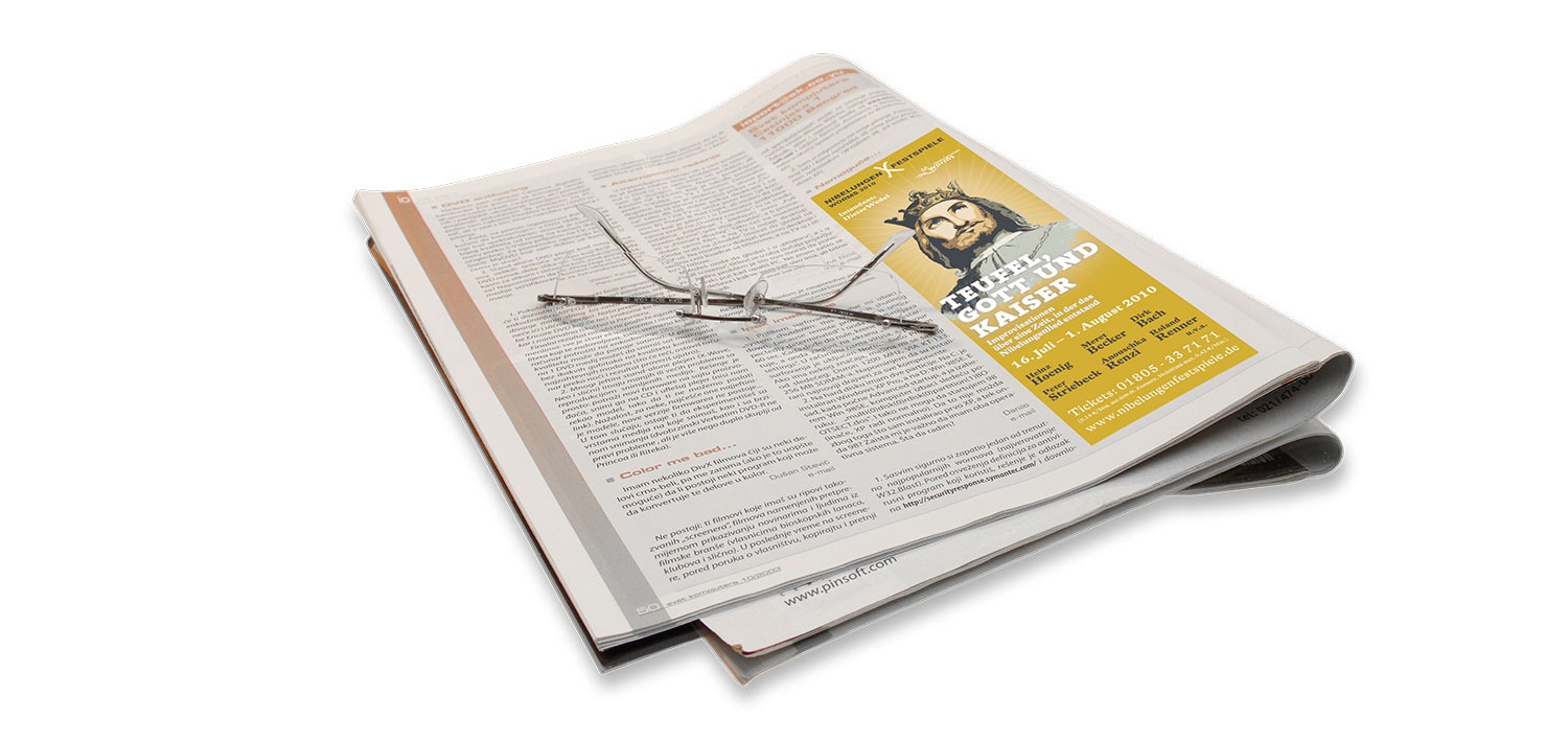 Eine silberne Lesebrille liegt auf einer aufgeschlagenen Zeitung mit Artikeln und einer gelben Anzeige der Vollblut-Nibelungen-Festspiele mit dem Porträt einer Person. Der Hintergrund ist weiß.