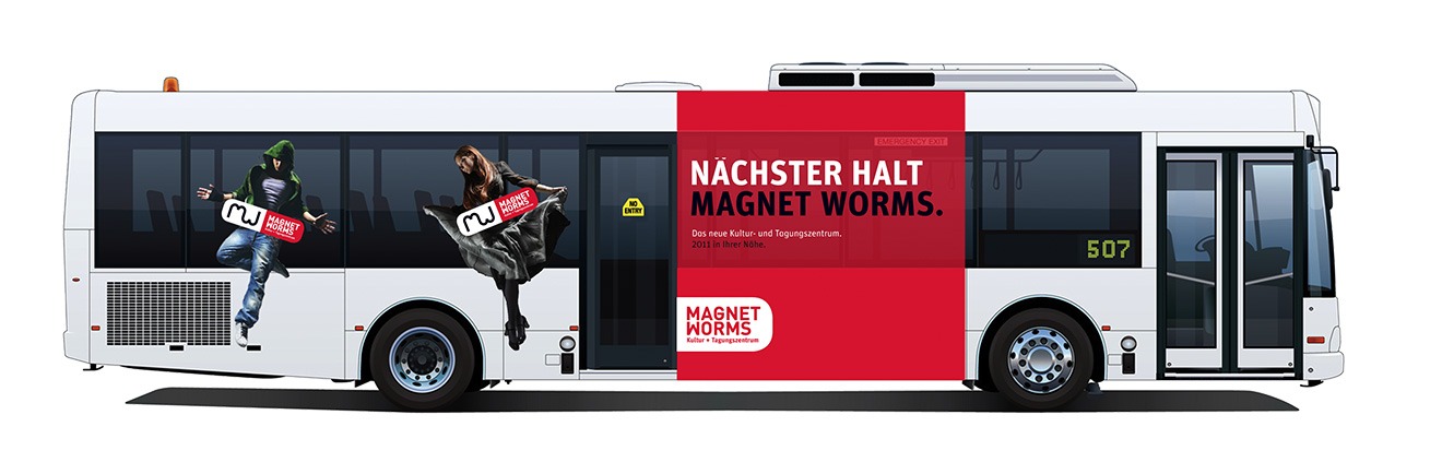 Ein weißer Stadtbus zeigt in kräftigen Rot-Schwarz-Farben Werbung für Magnet Worms. Die Werbung zeigt zwei Vollblut-Wormser, die mit Fahrkarten in der Hand springen. Der Text lautet: „Nächster Halt Magnet Worms“. Die Busnummer 507 ist deutlich zu erkennen.