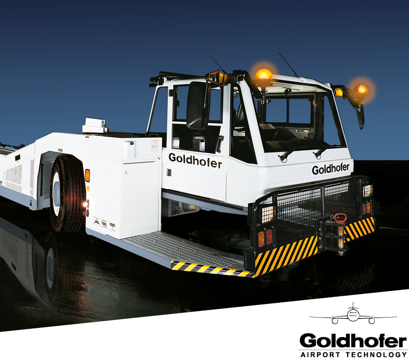 Ein weißer Goldhofer-Flughafenschlepper mit gelben Warnleuchten, schwarz-gelben Warnstreifen und dem Goldhofer-Logo steht auf einer dunklen, reflektierenden Fläche, in der Ecke das Firmenlogo